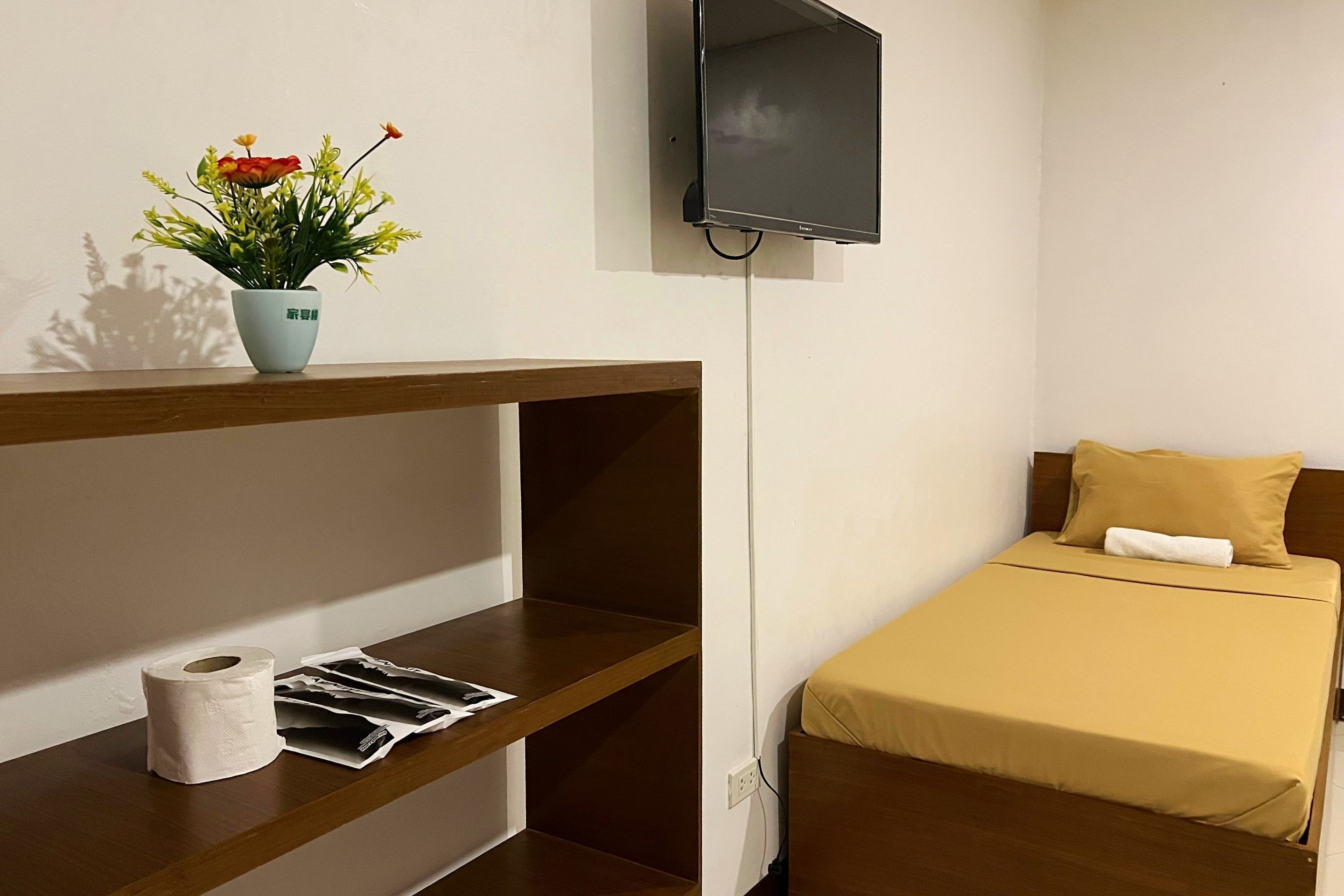 Quarto com cama, televisão na parede, prateleira de madeira com um vaso de flores e um rolo de papel higiênico.