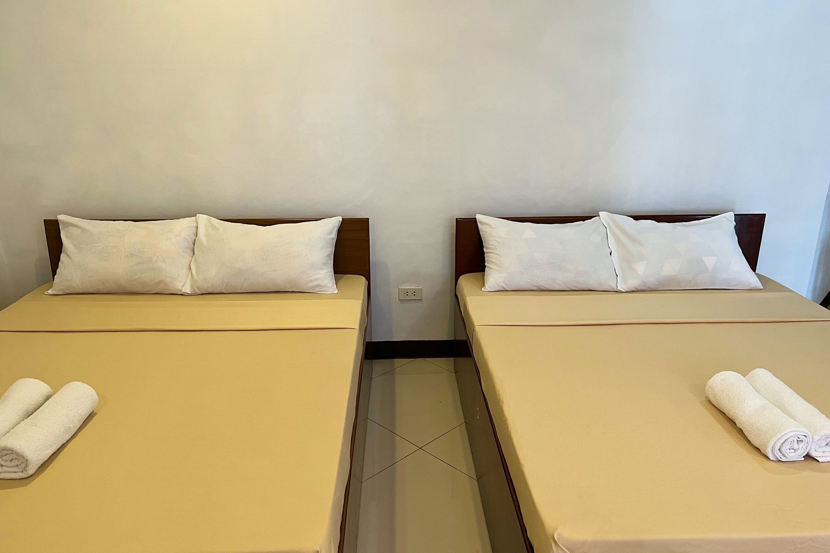 Deux lits simples avec du linge de lit et des oreillers de couleur beige, un sol carrelé et un mur uni.
