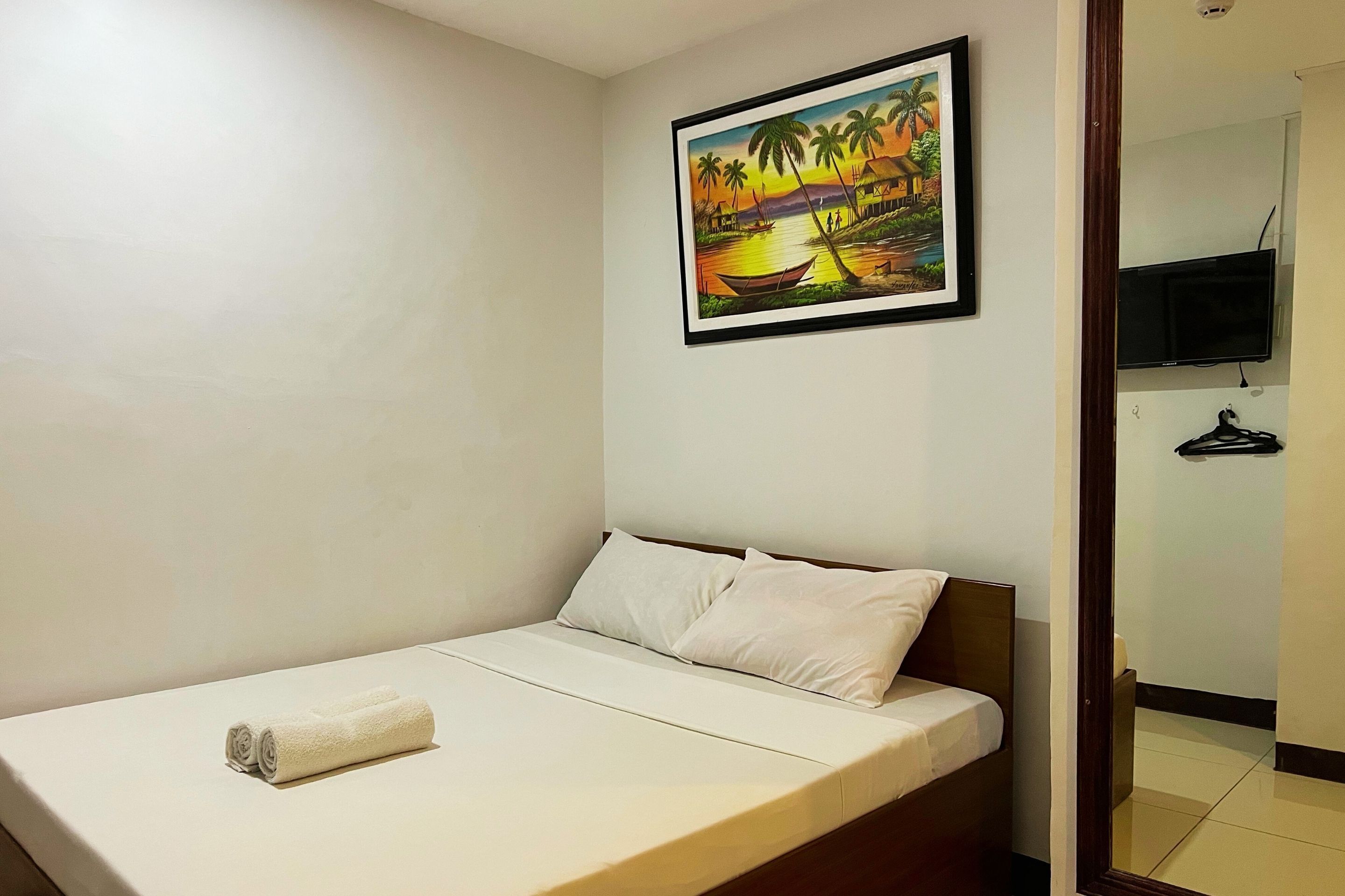Un lit simple avec un matelas et des oreillers blancs, un tableau représentant un paysage tropical accroché au mur et un téléviseur fixé au mur.