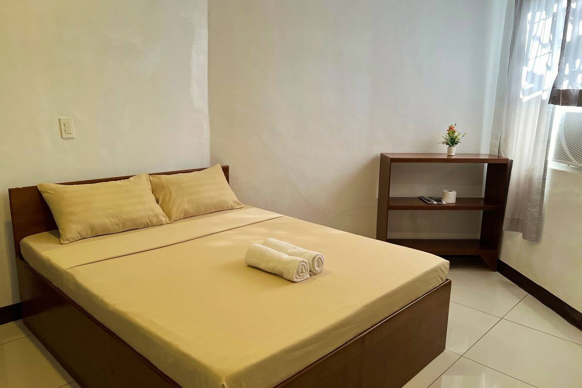 A neatly made bed with a beige bedspread and two pillows, a small wooden nightstand with a plant and a tissue box, and a window with sheer curtains.