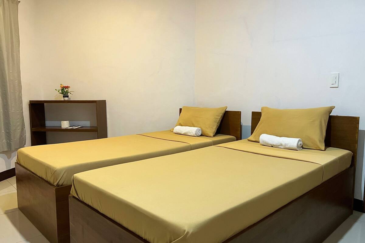 A room with two single beds, each with a wooden headboard and a yellow bedspread. There is a small wooden shelf with a vase of flowers and a book.