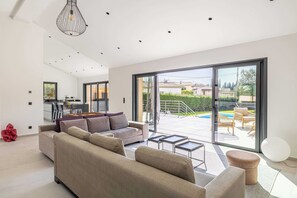 A modern living room with a sofa, coffee table, and a round ottoman. There is a dining area with a table and chairs, and a kitchen in the background. - Modern Villa With Heated Pool and Garden (Mougins)