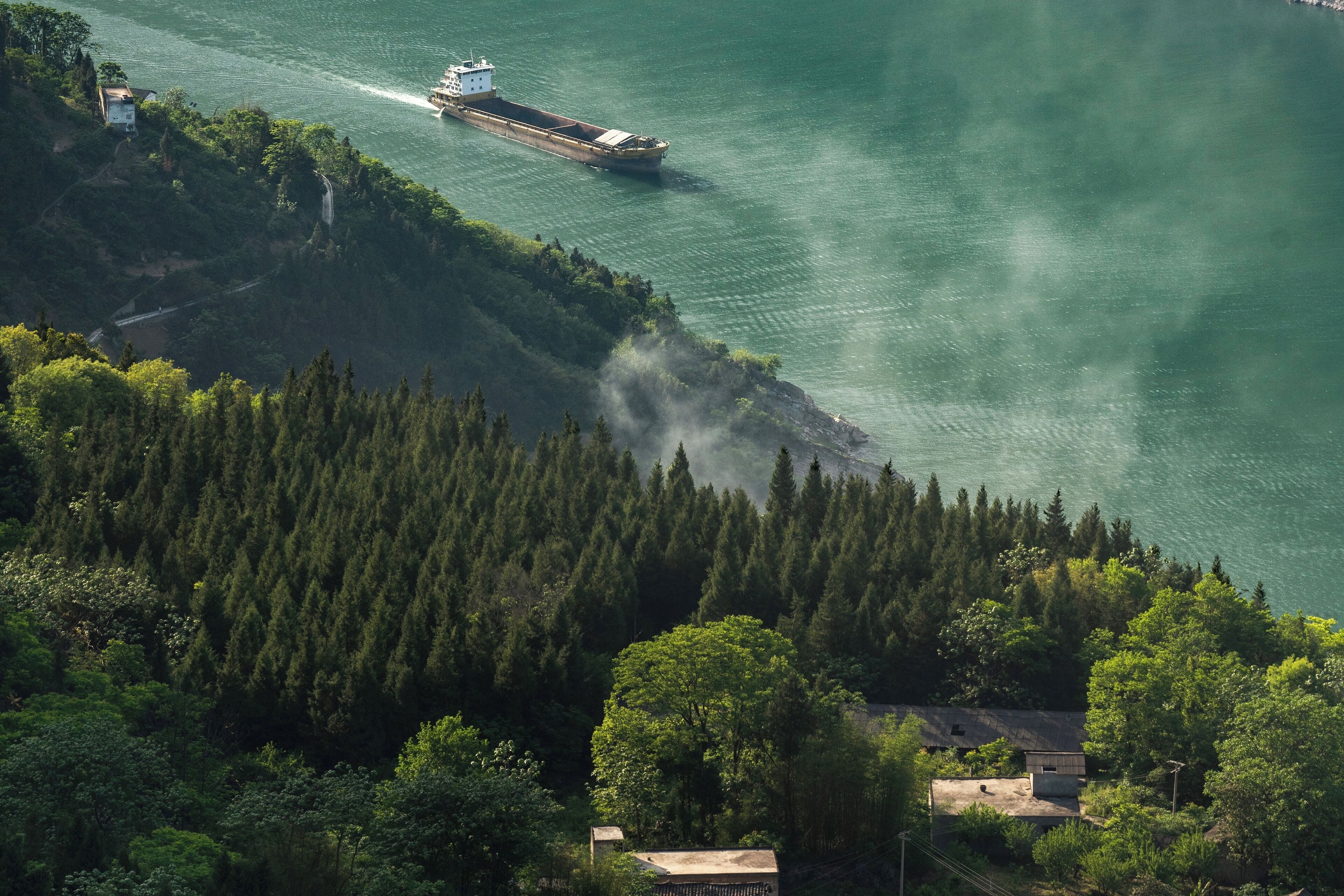 A large body of water with a boat, dense forest, and a few buildings on the shore.