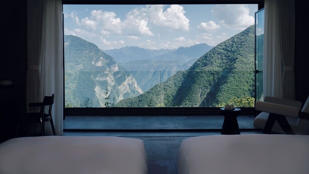 Ein Zimmer mit einem großen Fenster, das einen Blick auf eine gebirgige Landschaft freigibt. Es gibt zwei Betten und einen kleinen Tisch mit einer Tasse darauf.