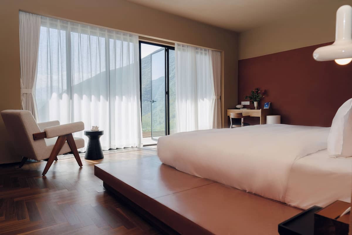 Moderni hotellihuone, jossa on suuri sänky, oleskelualue, jossa on tuoli ja pieni pöytä, sekä näkymä ulos liukuoven kautta.