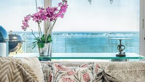 A modern living room with a large window overlooking the sea, a sofa with floral cushions, a glass coffee table, and a vase with pink orchids. - Stylish Camps Bay Home with Sea Views Beleza Villa (Cape Town)