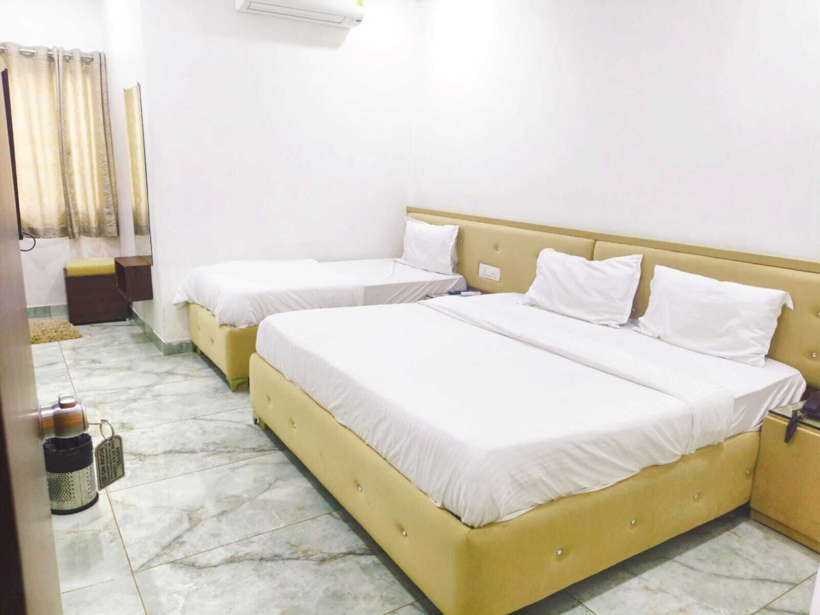 A hotel room with two beds, a small bedside table, a trash bin, and a window with curtains.