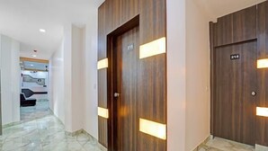Ein Korridor mit Holztüren, die mit 215 und 214 gekennzeichnet sind, Marmorboden und einem Einblick in einen modernen Innenraum mit einer Couch und einer Kaffeemaschine.