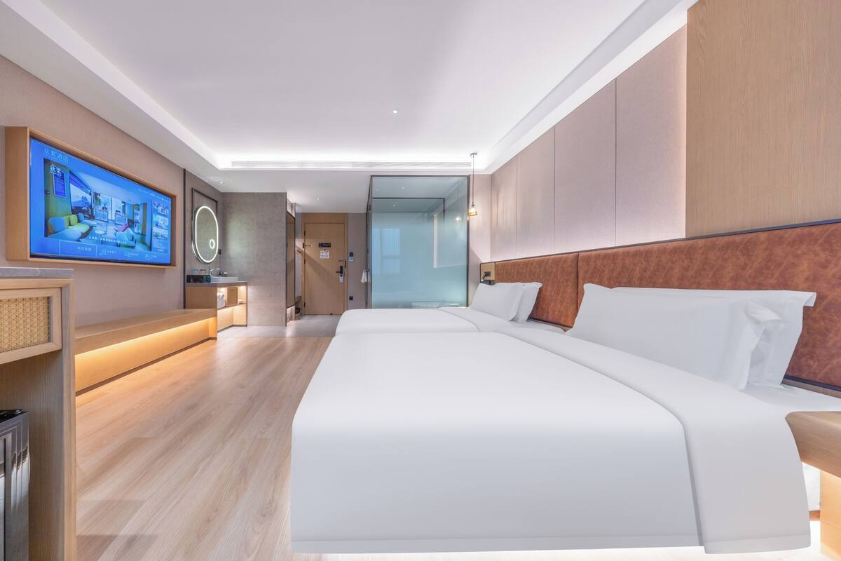 A modern hotel room with a large bed, a TV mounted on the wall, and a bathroom visible through a glass door.