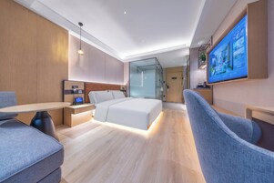 A modern hotel room with a bed, a desk, a chair, a TV, and a glass partition.