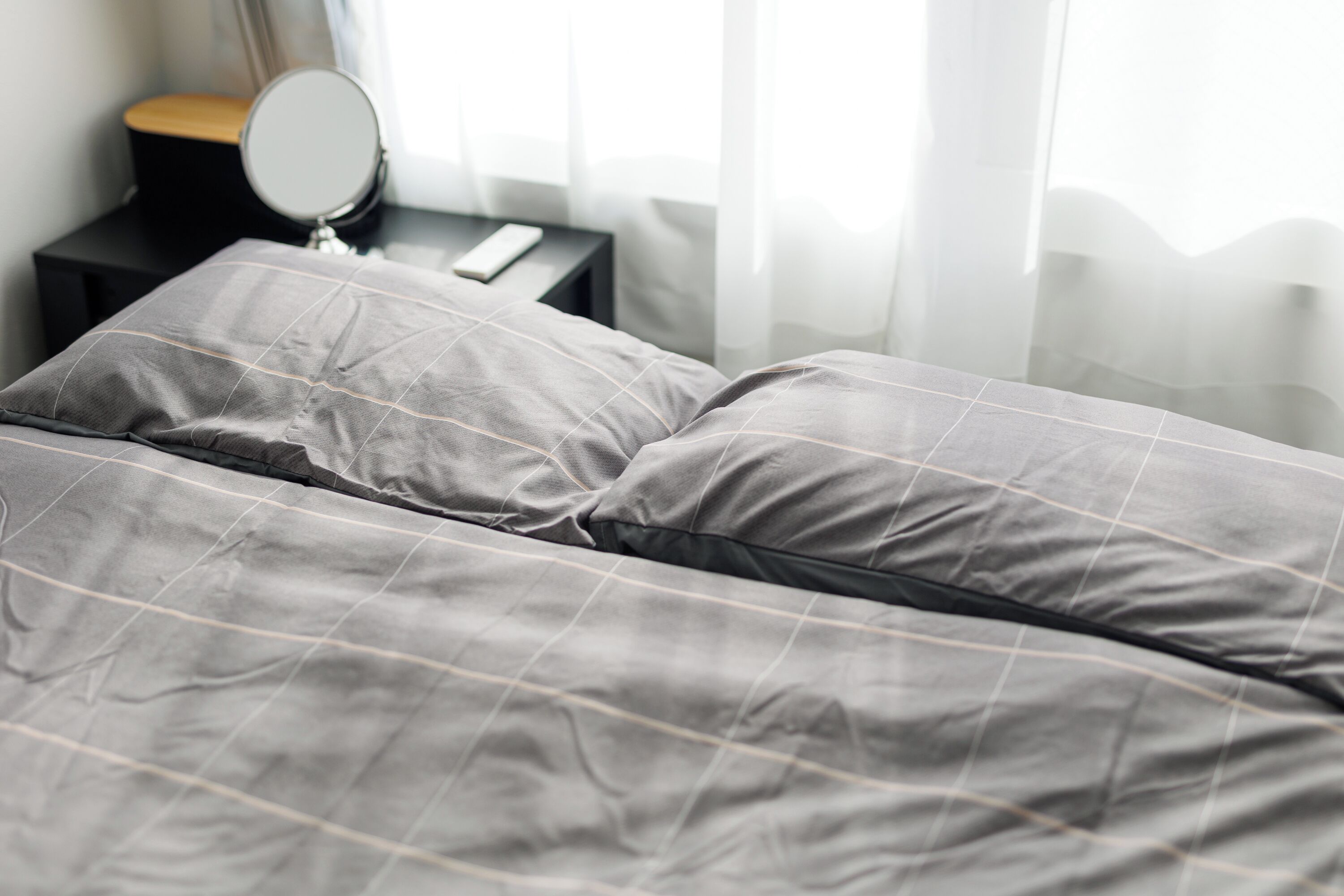 Una cama bien hecha con colcha gris y almohadas, una mesita de noche con espejo, y una ventana con cortinas translúcidas.