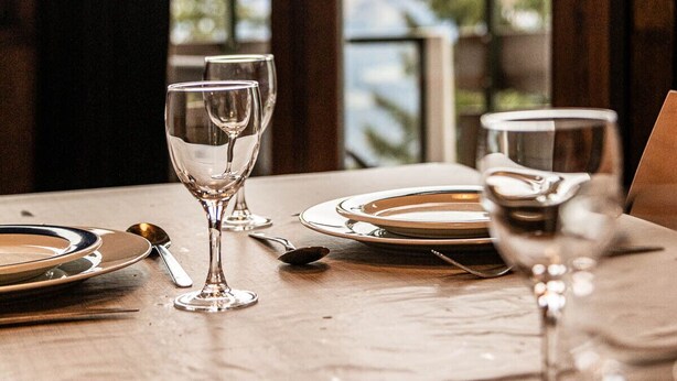 Une table dressée pour le dîner, avec des verres à vin et des assiettes, donnant sur un paysage magnifique à travers de grandes fenêtres.