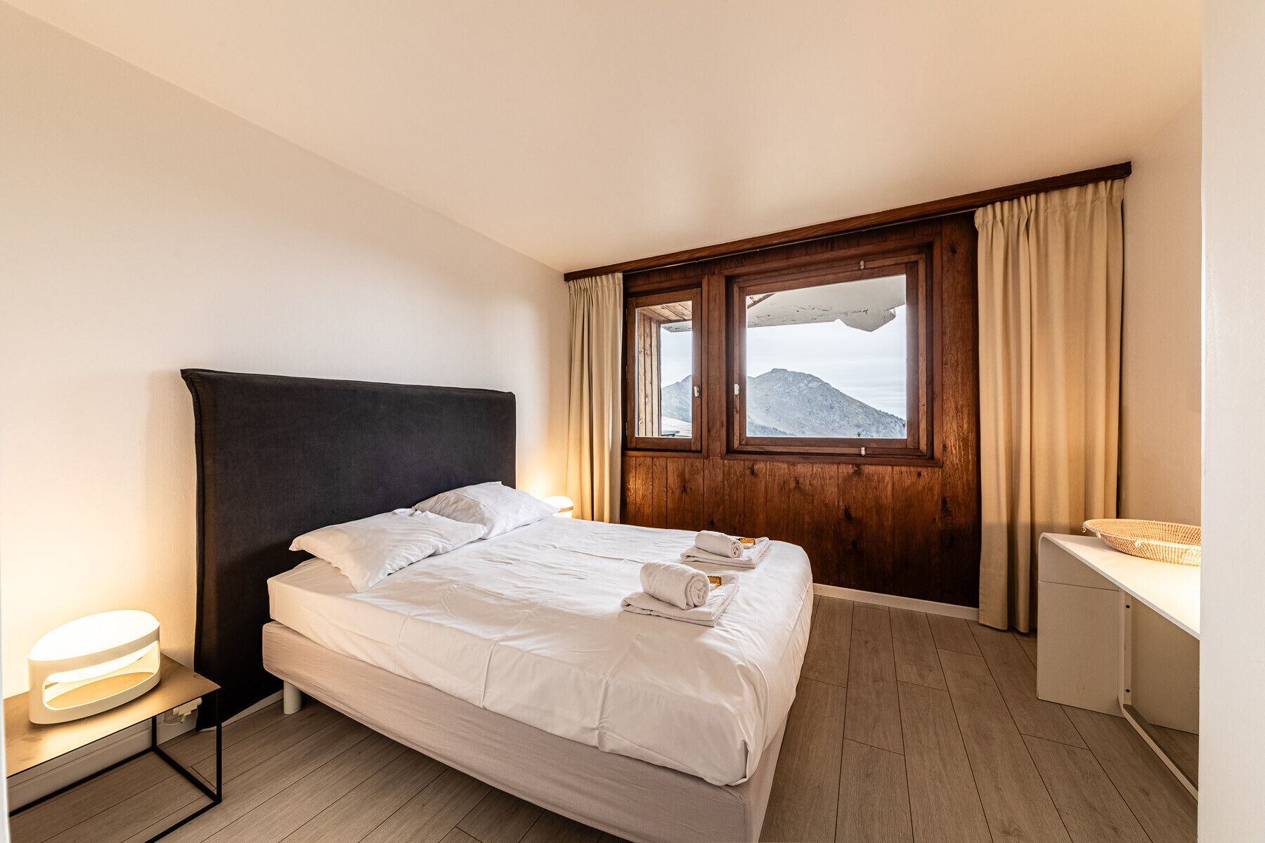 A hotel room with a bed, a wooden headboard, a window with a mountain view, a bedside table, and a small desk with a lamp.