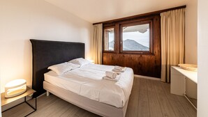 Une chambre d’hôtel avec un lit, une tête de lit en bois, une fenêtre donnant sur les montagnes, une table de chevet et un petit bureau avec une lampe.