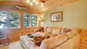 A cozy cabin interior with a wooden ceiling, a large window, a cushioned sofa, and a framed picture on the wall. - Dock + Fire Pit: ‘Sweet Serenity’ on Amber Lake (Lac Du Flambeau)