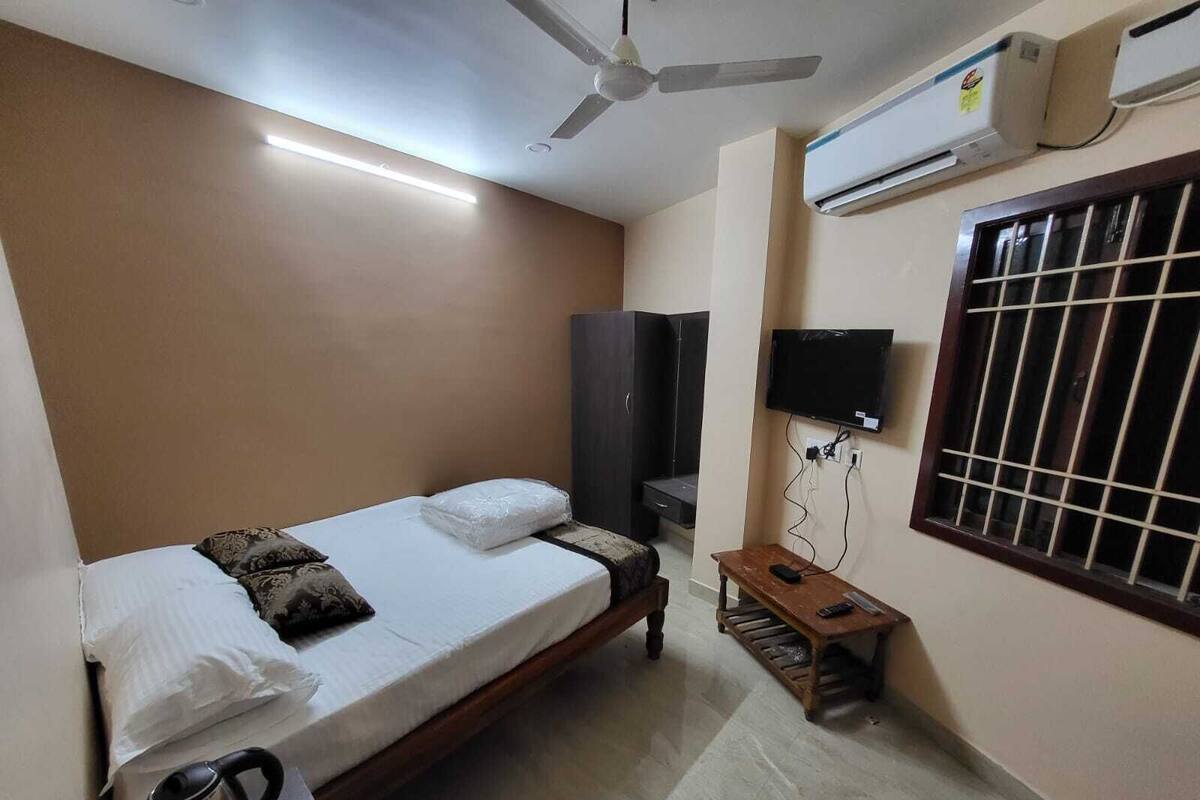 Una cama individual con almohadas, un ventilador de techo, un aire acondicionado, un televisor y una ventana con barrotes.