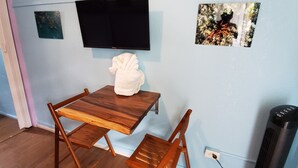 Una habitación con una mesa de madera y dos sillas, una toalla blanca sobre la mesa, un televisor de pantalla plana en la pared y un calentador en el suelo.