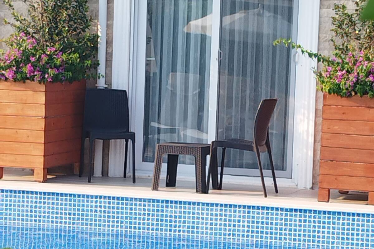 A swimming pool with two chairs and a small table, surrounded by wooden planters with flowers, against a stone wall with a sliding glass door.
