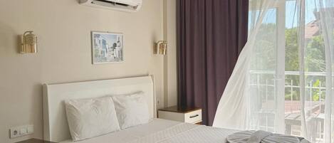 A hotel room with a bed, a nightstand, an air conditioning unit, a picture on the wall, and a window with curtains.