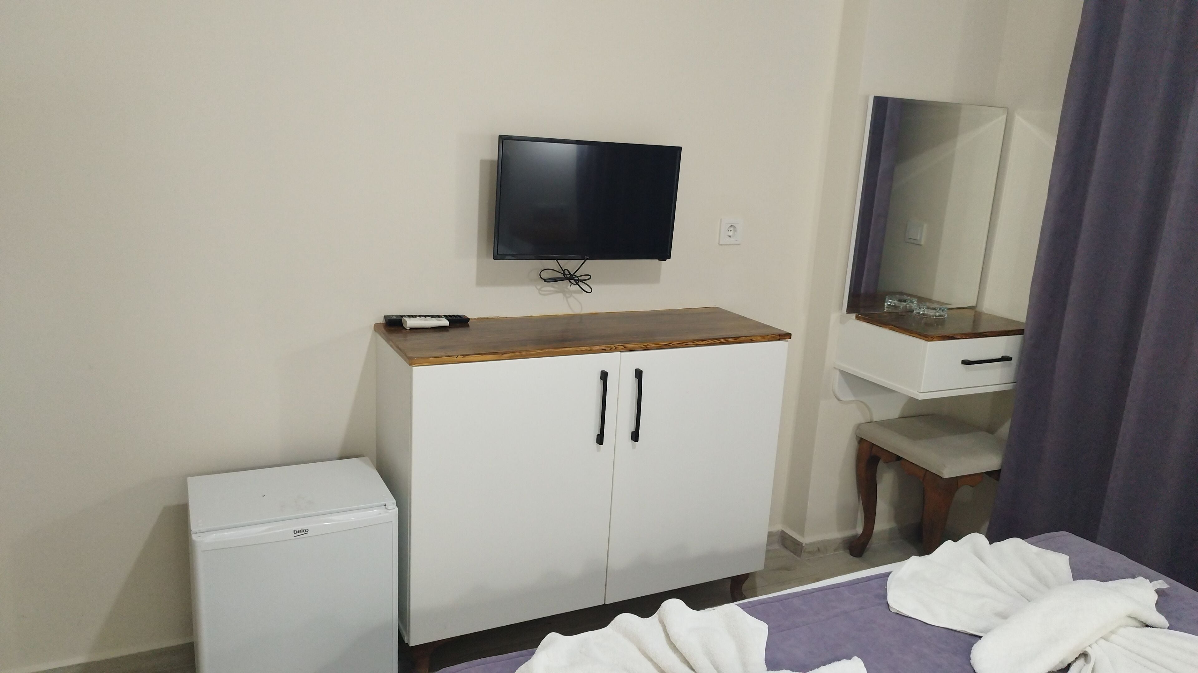 A hotel room with a bed, a small white refrigerator, a wooden cabinet with two doors, a television mounted on the wall, a mirror, and a window with curtains.