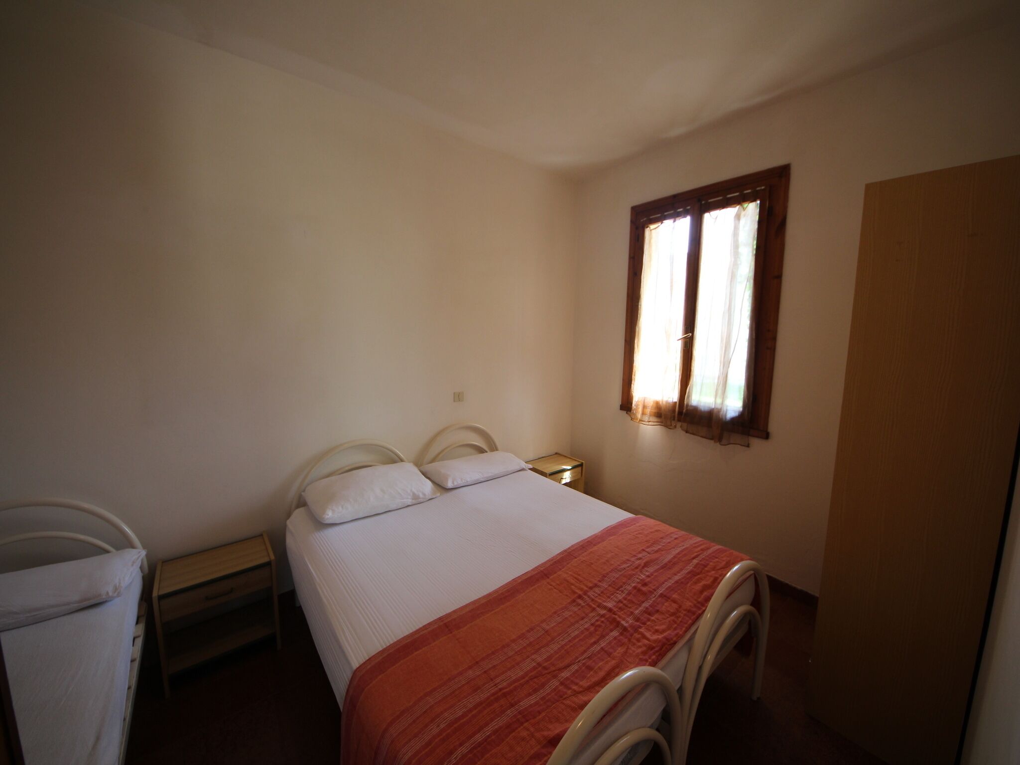 Quarto pequeno e simples com uma cama de solteiro, mesa de cabeceira, janela com cortinas e porta.