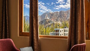 Une pièce avec un ordinateur portable posé sur une petite table, deux chaises rouges et une fenêtre donnant sur une montagne et un bâtiment.