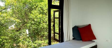 Una cama individual con un colchón blanco y dos almohadas, una roja y otra azul, en una habitación con un gran ventanal que da a una densa vegetación verde.