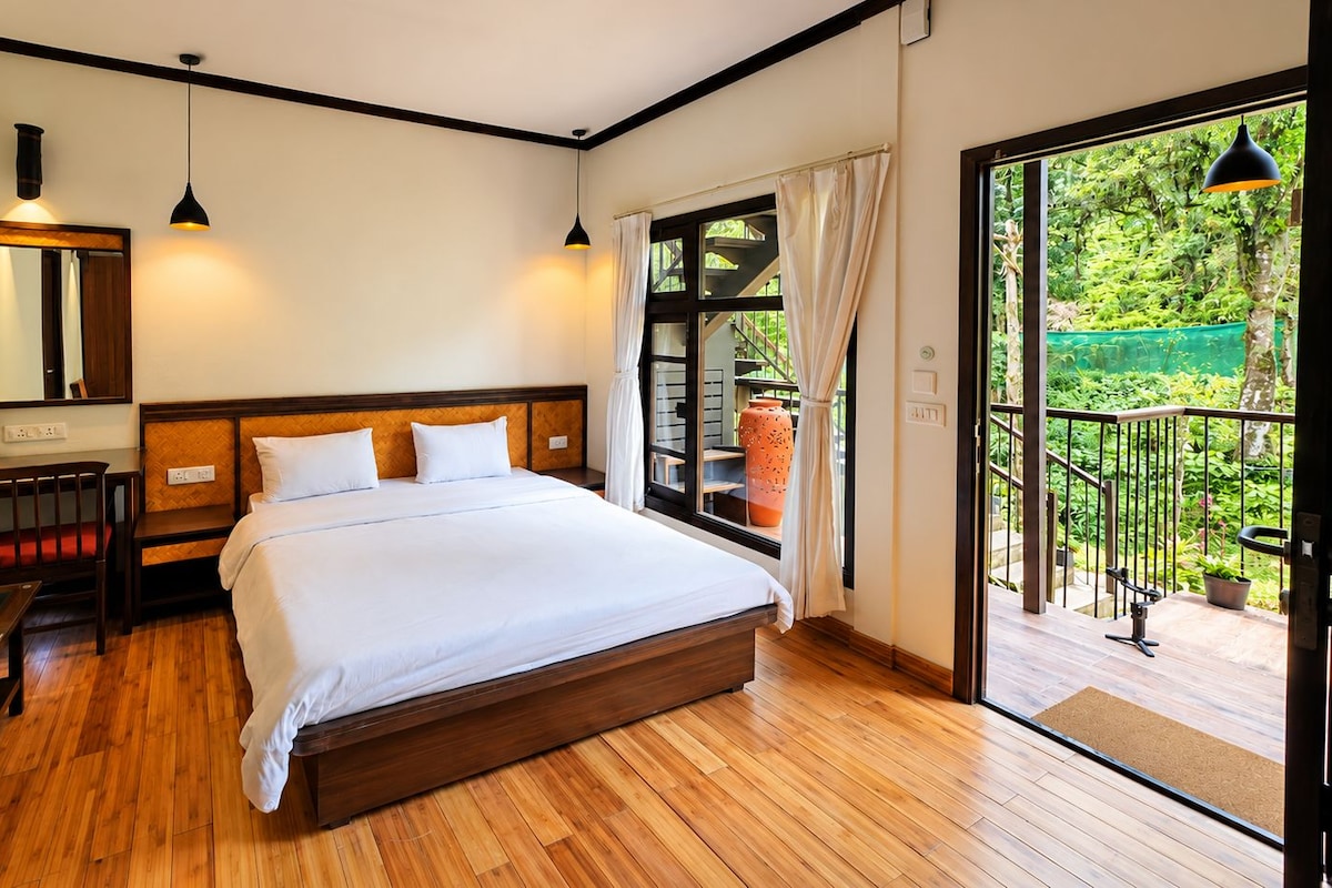A bedroom with a large bed, wooden flooring, a desk, a chair, a mirror, and a window with a view of greenery.