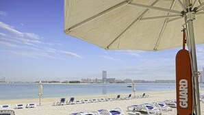 Une plage bordée de rangées de chaises longues et de parasols, un poste de surveillance pour les sauveteurs, et, au loin, le profil de la ville.