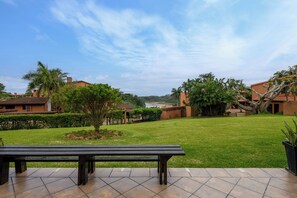 A patio area with a bench, a well-maintained lawn, and a view of a river and buildings in the distance. - San Lameer Villa 2006 (Southbroom)