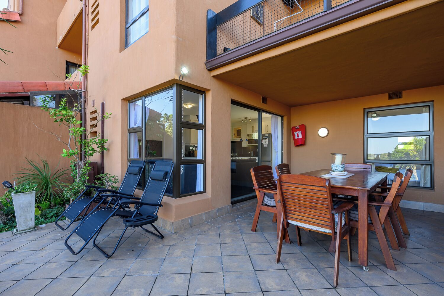 An outdoor patio with a dining table and chairs, a small garden with plants, and a building with large windows and a balcony.