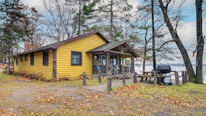 Keltainen mökki, jossa on kuisti, piknikpöytä ja grilli, ja se sijaitsee järven rannalla.
