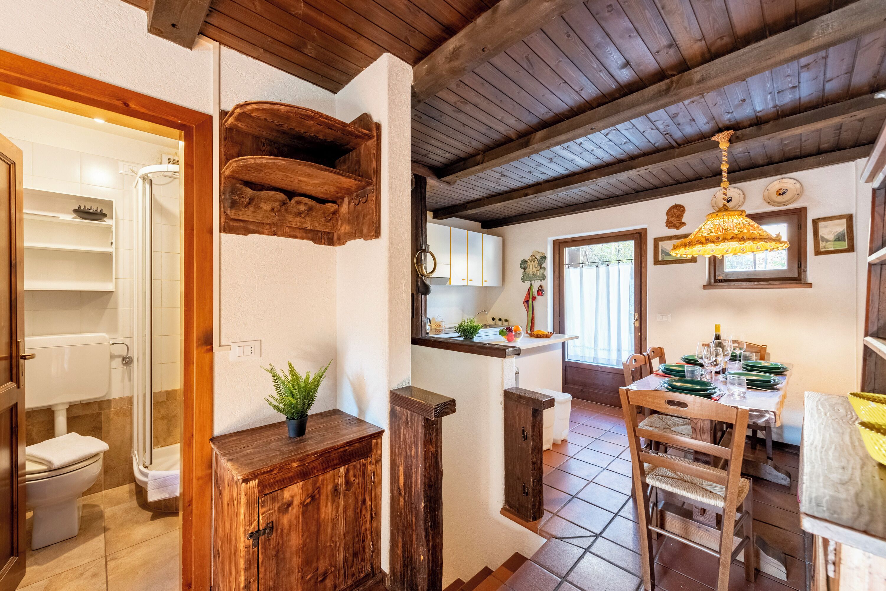 Un intérieur rustique avec un plafond en bois, un coin repas avec une table dressée pour quatre personnes et un coin cuisine équipé d’un évier et de placards.