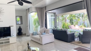 A modern living room with a flat-screen TV, a ceiling fan, a Buddha statue, a sofa with cushions, and a view of a pool and greenery outside.