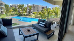 Ein Balkon mit einer Couch, einem Holztisch und einem Esstisch mit Stühlen. Im Hintergrund ist ein Swimmingpool zu sehen.