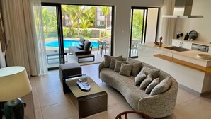 Ein modernes Wohnzimmer mit Sofa, Couchtisch und Essbereich. Eine Küche ist sichtbar, mit Spüle und Arbeitsplatte. Große Fenster bieten Blick auf einen Swimmingpool und Palmen.