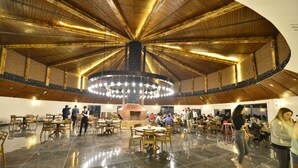 A spacious indoor dining area with a high ceiling and a central circular chandelier. Tables are arranged around a central fireplace. - Kayı Resort Hotel (Sarikamis)