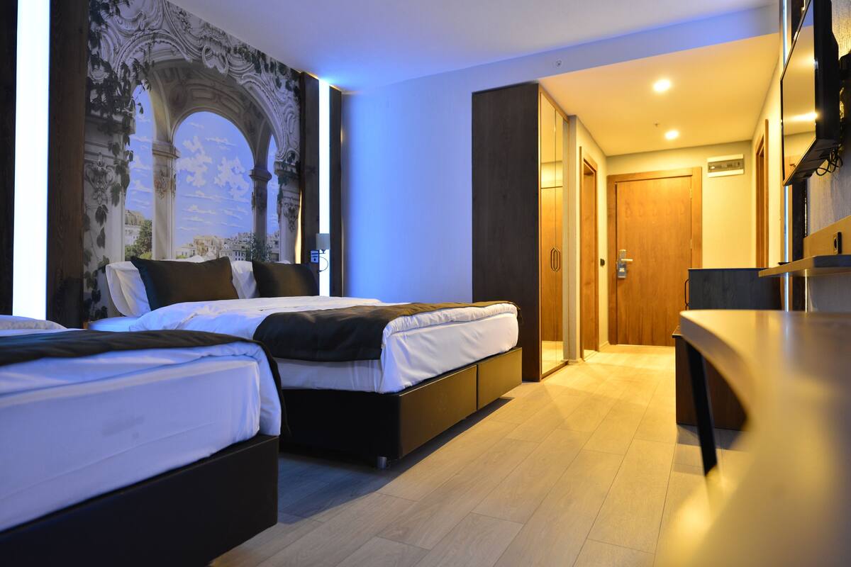 A modern hotel room with two beds, a dining area, and a large mural on the wall.