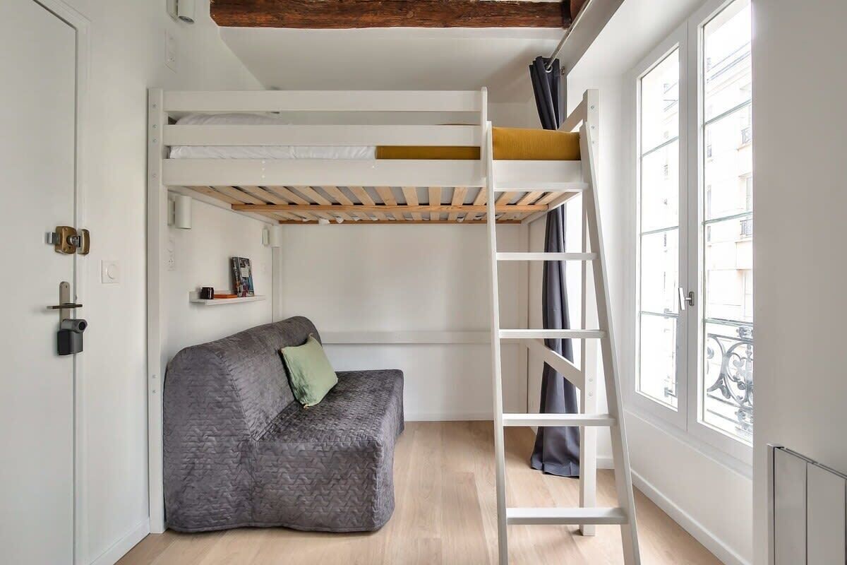 A compact room with a bunk bed, a ladder, a gray sofa, and a window with a view of buildings.