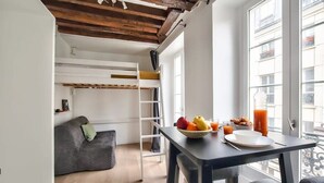 A compact room with a bunk bed, a small sofa, a dining table with breakfast items, and a window with a view of buildings. - Cosy Mezzanine Studio in the Heart of Paris (4) (Paris)