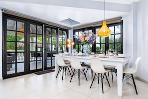 A modern dining area with a white table set for four, surrounded by white chairs with black legs. A large vase with flowers is on the table, and there are two hanging pendant lights above.