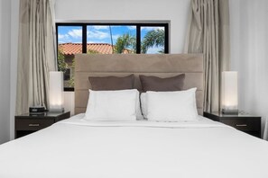 Une chambre d’hôtel avec un grand lit, deux tables de chevet, une fenêtre donnant sur des palmiers et un immeuble, ainsi que des rideaux.
