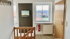A room with a wooden chair, a framed painting, a window with a view of houses, a radiator, and a fire extinguisher.