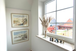 A room with two framed paintings on the wall, a window with a view of a building, and a shelf with a vase and decorative letters spelling 'BEACH'.