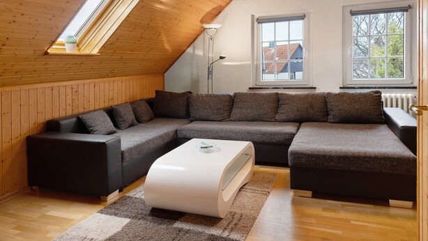 Un moderno espacio tipo loft con sofá seccional, una mesa de centro blanca, piso de madera y un techo inclinado con tragaluces.