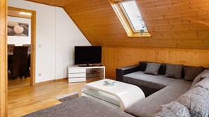 Una habitación moderna estilo loft con techo inclinado, un televisor sobre una consola blanca, un sofá seccional gris y un tragaluz.