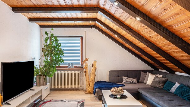 Un salón estilo loft con techo inclinado, televisor de pantalla plana, sofá gris, mesa de centro y un amplio ventanal con persianas.