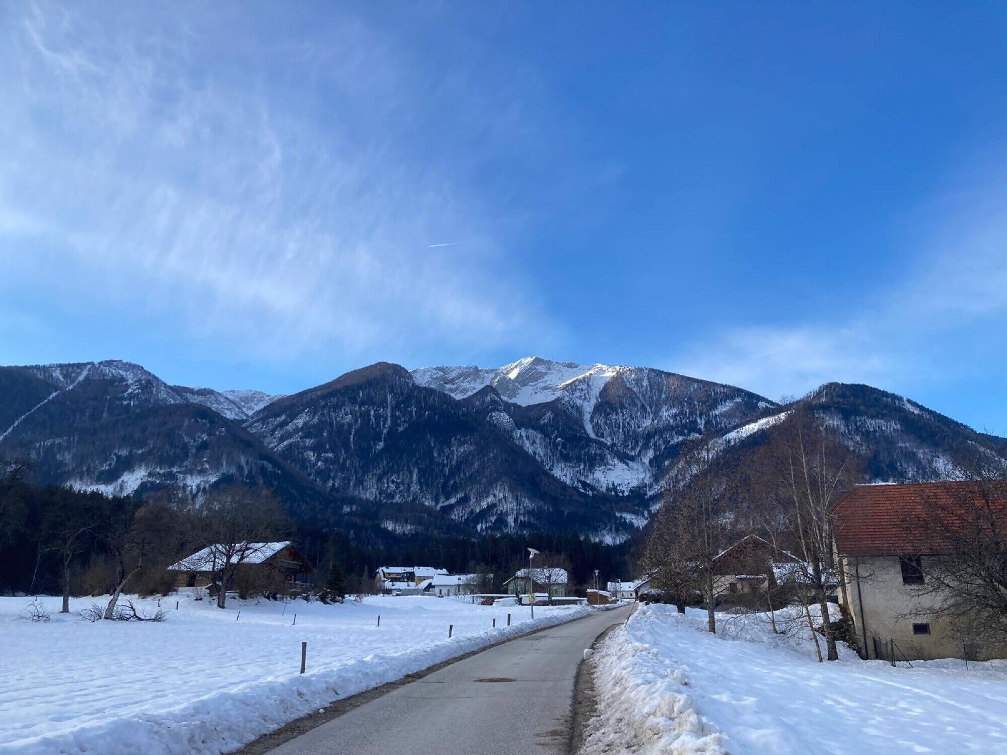 Une route de campagne enneigée, bordée de maisons de chaque côté, avec des montagnes en arrière-plan et un ciel dégagé.