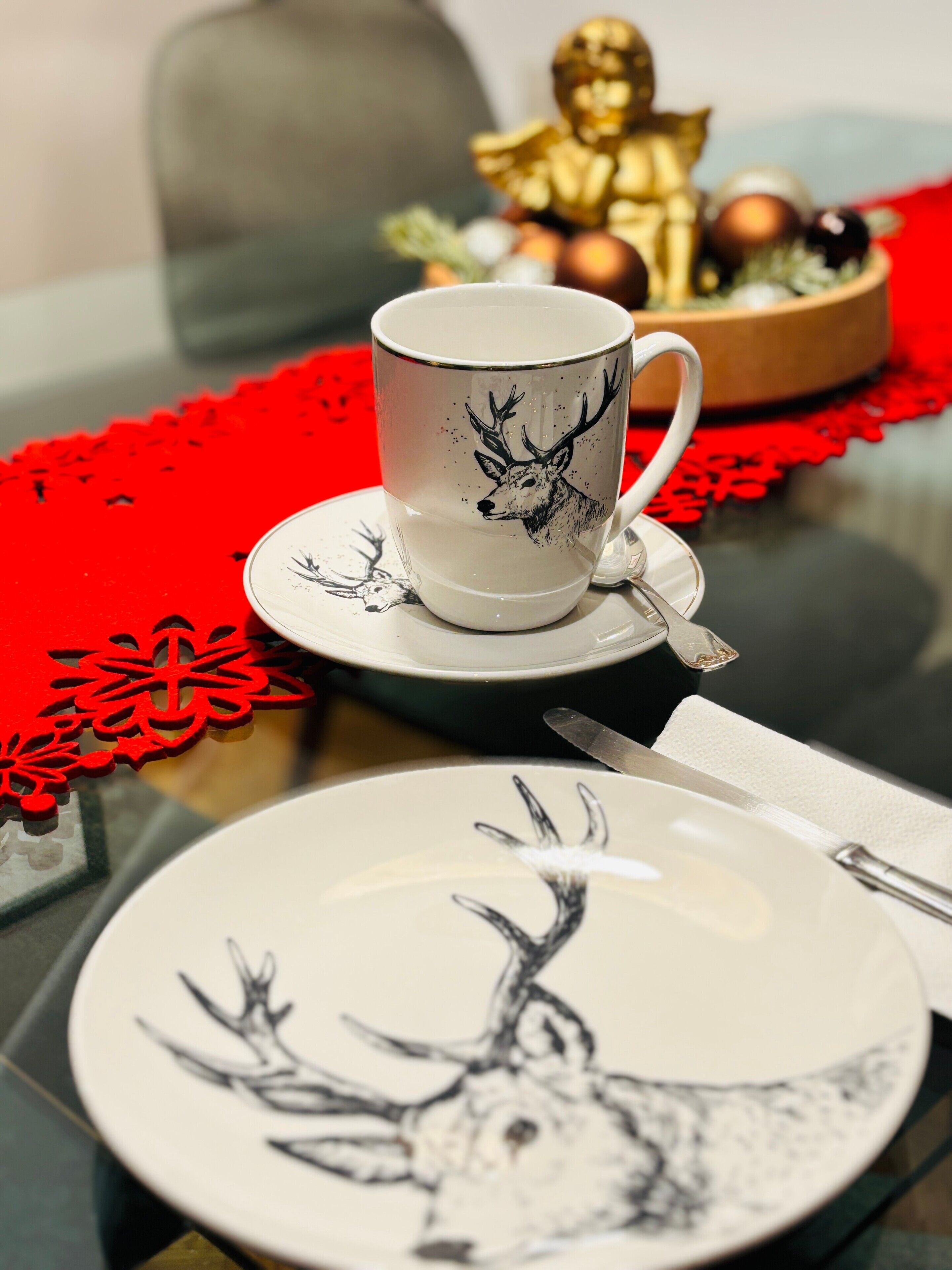 Ein Tisch mit einer weißen Tasse mit einem Hirsch-Motiv, einem weißen Teller, der ebenfalls einen Hirsch zeigt, einer goldenen Figur und einem roten Dekorationsgegenstand.