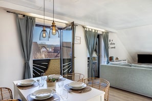 Un coin repas avec une table dressée pour quatre personnes, un canapé et un téléviseur, dans un appartement moderne doté de grandes fenêtres et de rideaux.